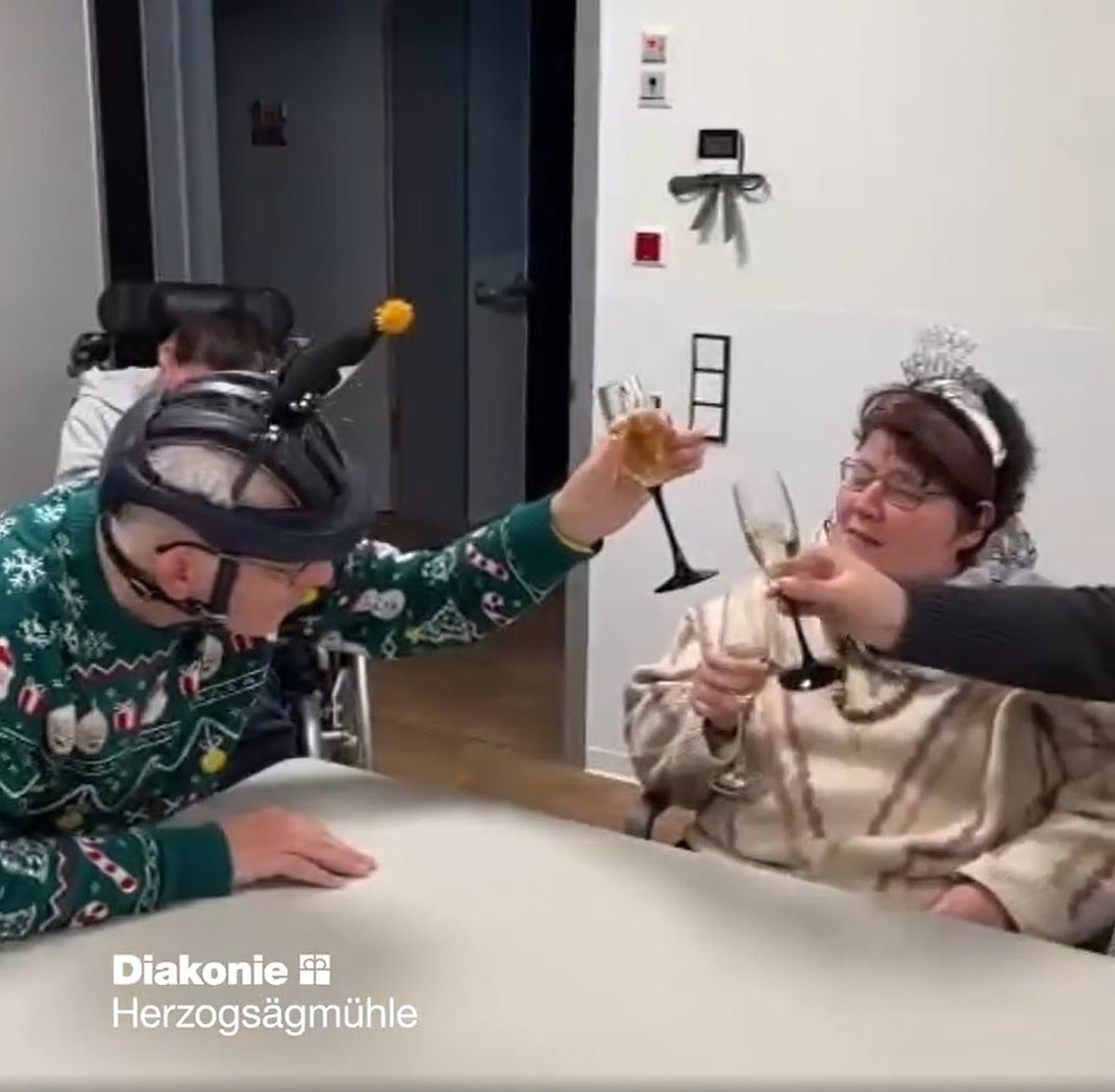 Zwei Feiernde mit Behinderung am Tisch und eine weitere Person stoßen mit Sektgläsern an.