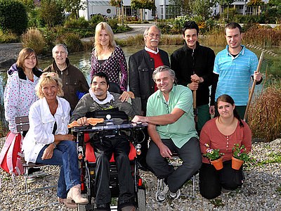 Zehn Personen, unterschiedlichen Alters, mit und ohne Behinderung, stehen und sitzen als Gruppe vor einem Teich.