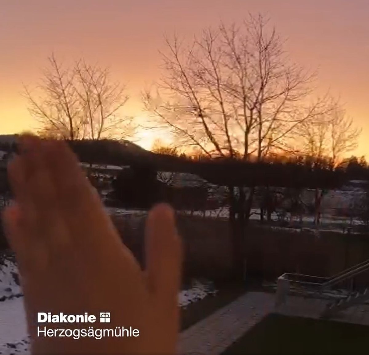 Blick in die Winterlandschaft mit untergehender Sonne. Im Vordergrund ist eine winkende Hand zu sehen.