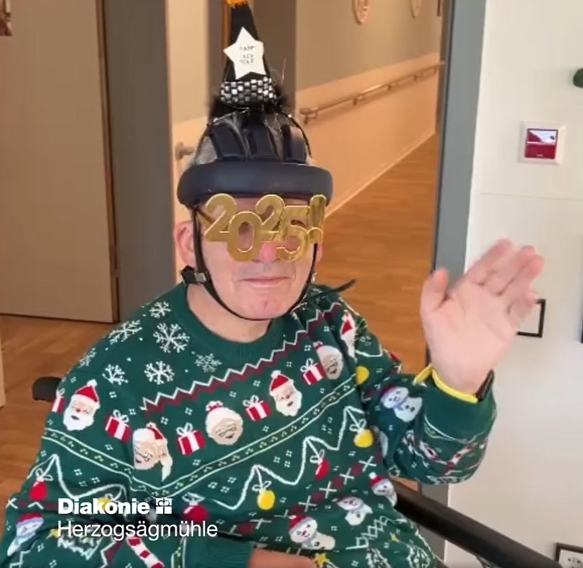 Eine sitzende Person mit Weihnachtspullover winkt in die Kamera. Eine goldene 2025 ist als Brille ins Bild montiert.