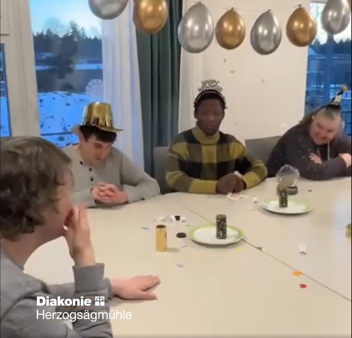 Feiernde am Tisch. Der Raum ist mit einer Kette aus goldenen und silbernen Luftballons geschmückt.
