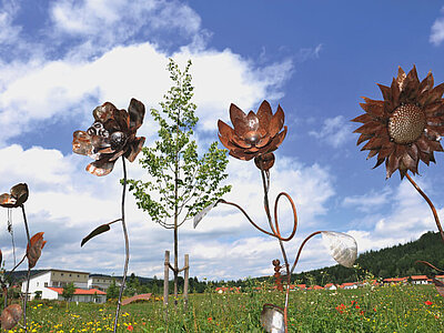Mehrere handwerklich gefertigte Sonnenblumen aus Metall auf einer Blumenwiese mit blauem Himmel und weißen Wolken im Hintergrund