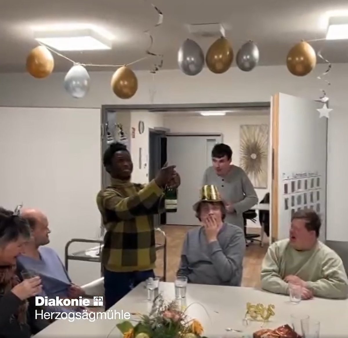 Vier Feiernde sitzen am Tisch, eine Person steht, eine kommt gerade dazu.