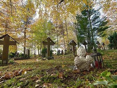 Detail-Aufnahme des Friedhofes Herzogsägmühle; inmitten eines Waldes stehen Grabkreuze, davor ein Stein-Engel mit einer Grab-Laterne.