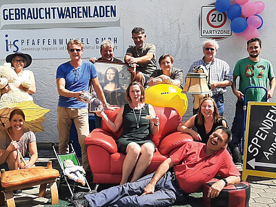 Mehrere Menschen lachend mit verschiedenen Artikeln des Gebrauchtwarenladens