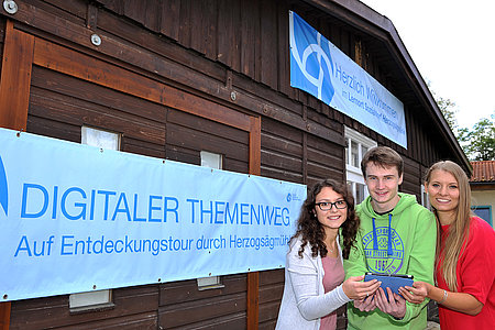 Drei junge Menschen halten ein Tablet für den Besuch der digitalen Themenwege in der Hand und schauen frölich in die Kamera.